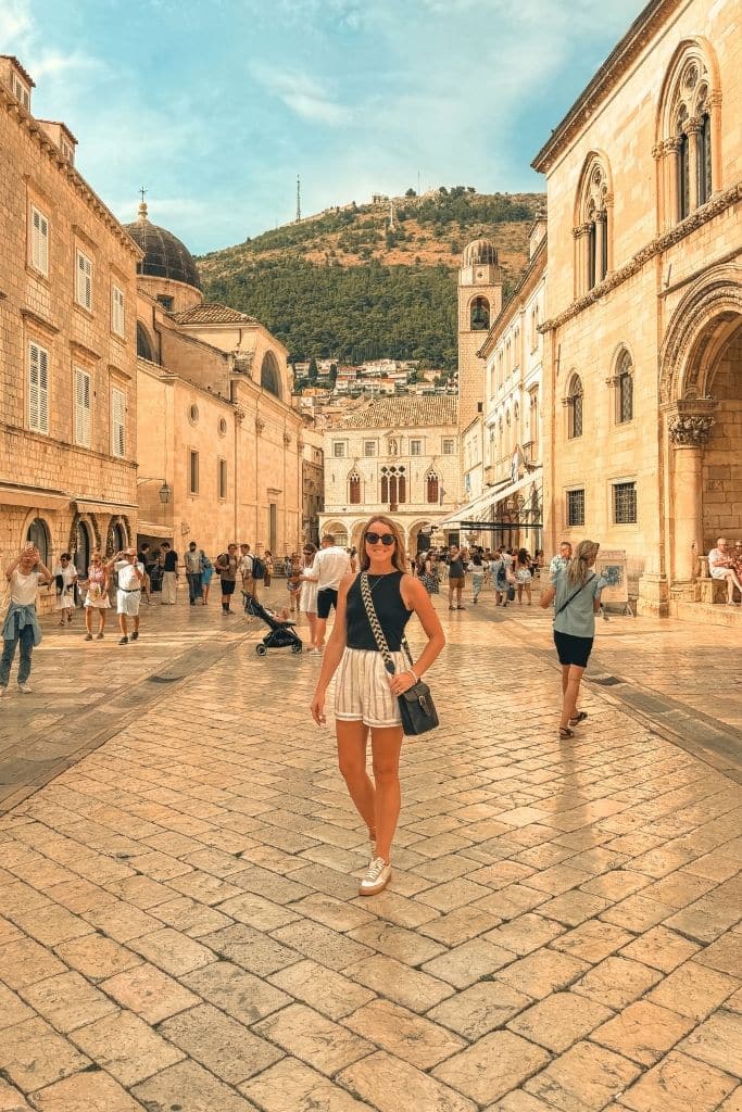 Kate wearing a simple black tank top, lightweight striped shorts, comfortable sneakers, and a crossbody bag while exploring Europe’s cobblestone streets in summer.