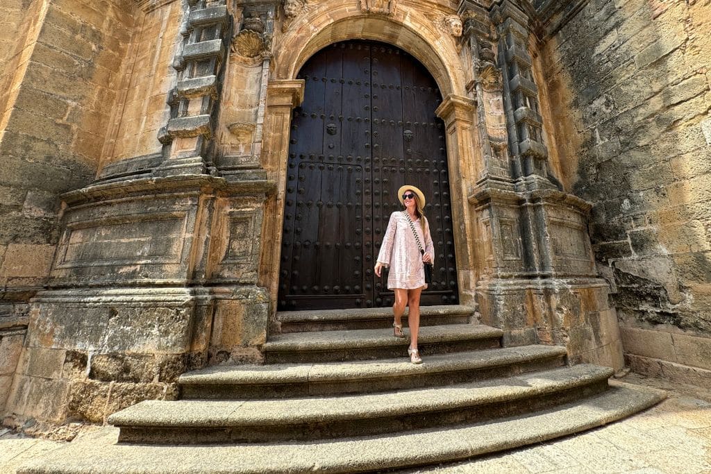 Kate explores Europe in summer standing beneath a massive historic doorway, surrounded by beautiful old architecture and warm Mediterranean sunshine.