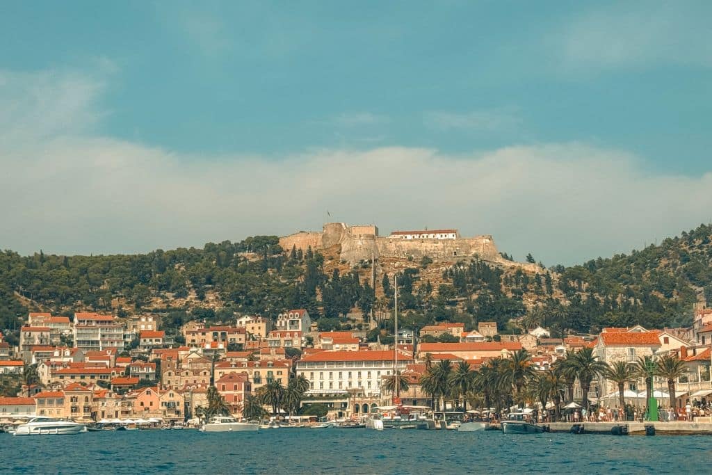 Stunning shoreline of Hvar Island in Croatia, lined with old stone buildings, palm trees and an old fort perched on the hill over the old town.