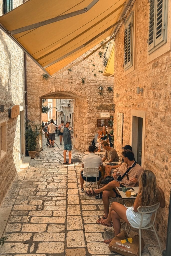 Narrow alley in Hvar with a cafe and people sitting outside at small tables.