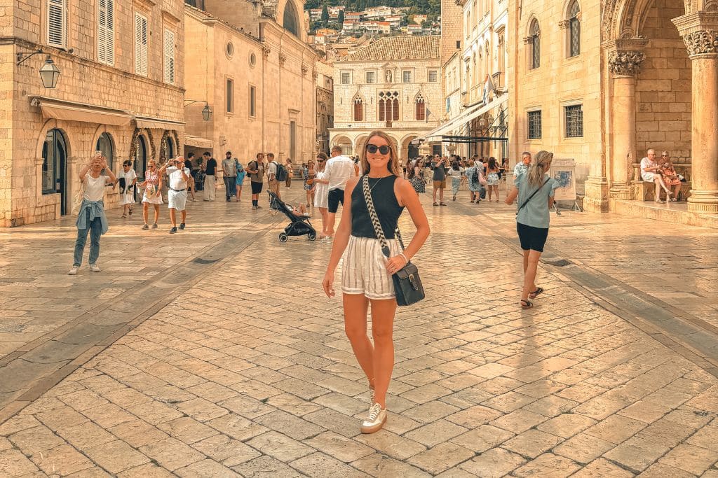 Kate smiling at the camera, standing on a cobble stone street in Dubrovnik, wearing linnen shorts and a black tank top with a crossbody bag.