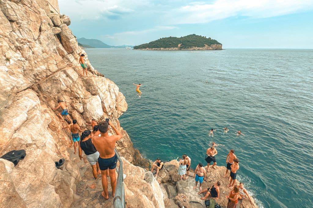 People in swimwear standing on steep rocky cliffs as someone is jumping into the water below.