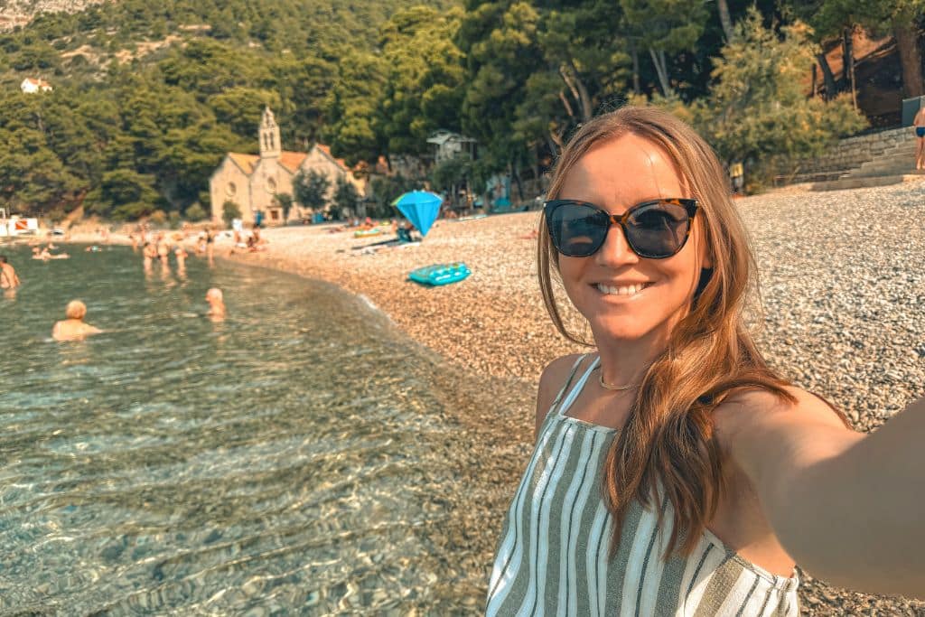 Kate smiling as she takes a selfie standing on a pebble beach with an old church in the background in Croatia.