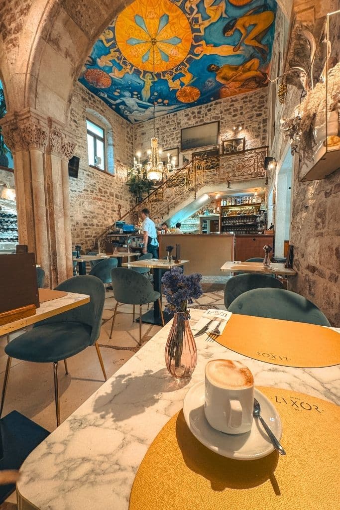 Stone walls and frescos on the ceiling inside Lvxor Restaurant in Split with a Cappuccino on the yellow tablecloth.