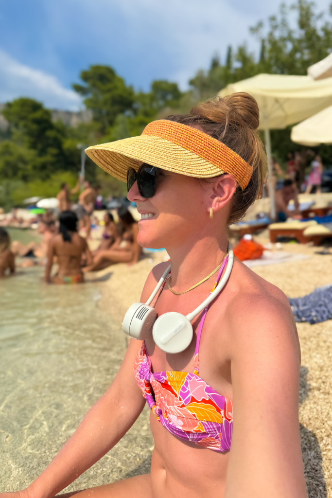 Kate is sitting in the water on the edge of a beach with a sunhat and a neck fan around her neck, smiling out at the water.