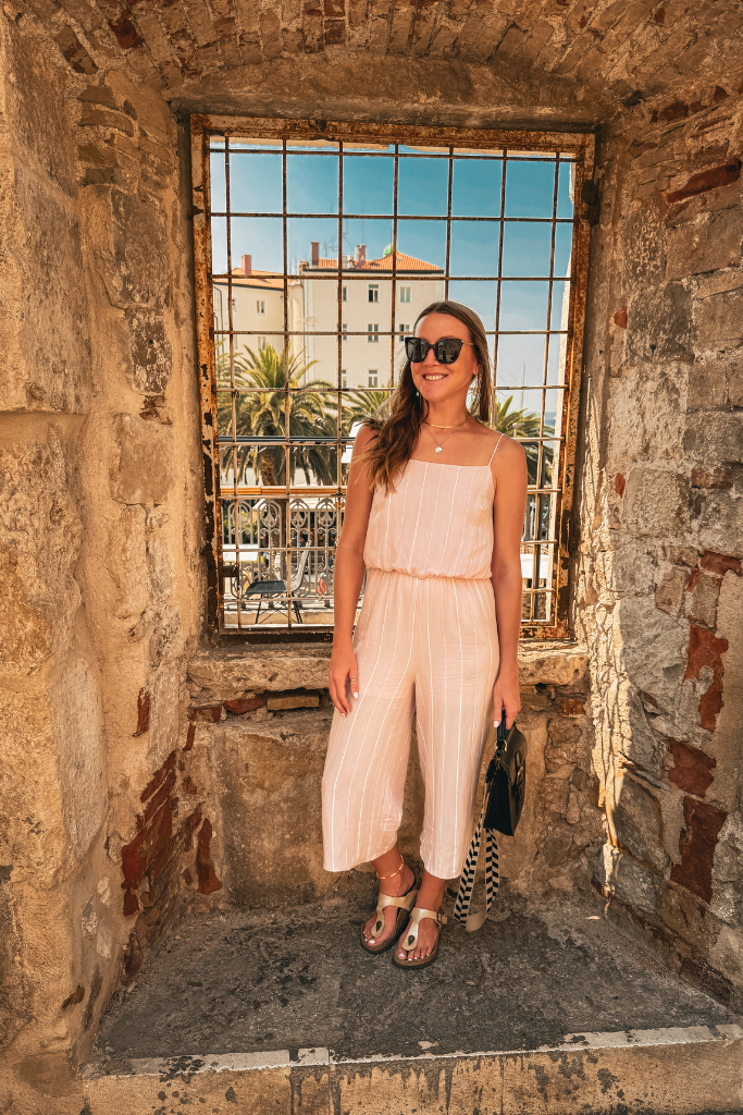 Kate is standing in a window nook in the old wall surrounding Split. A view of palm trees through the window.