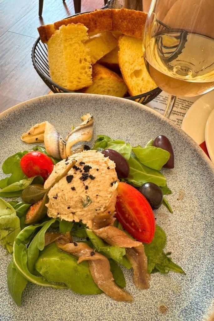 Plate of fresh sardines and salad with a basket of bread and a glass of white wine.
