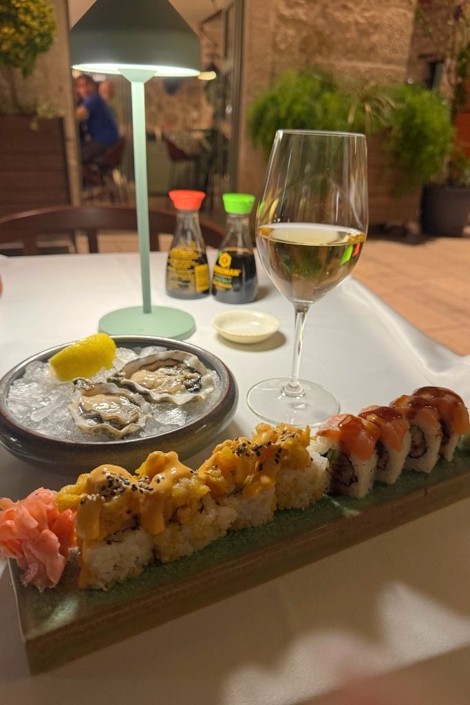 A sushi roll and two oysters with lemon and a glass of white wine on a white table cloth in the evening lit by a small table lamp.