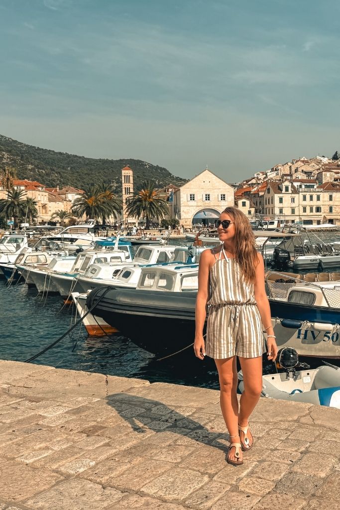 Kate standing on walkway with the marina and old town of Hvar behind her. It's a sunny day and she's wearing a short romper.