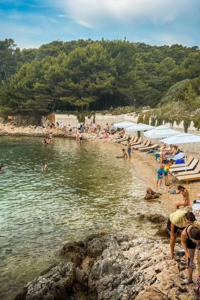 A small crescent-shaped beach with beach chairs and umbrellas lined up all along it. The shore is rocky but the water is clear.