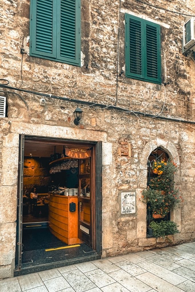 Store front of a cafe in an old stone building on a cobble stone street with green shutters and flower pots hanging.