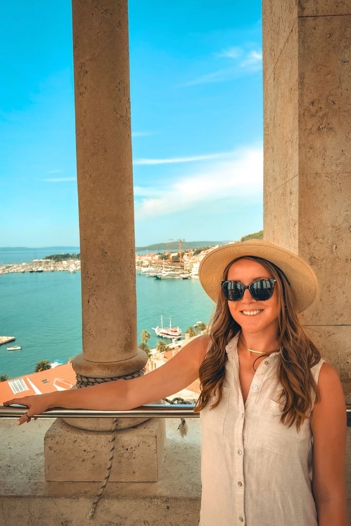 Kate leans one arm over a railing at the top of a tower with views of Split's marina through the narrow window.