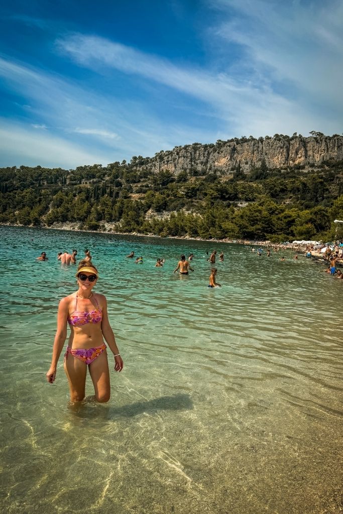 Kate wades out knee deep in the crystal clear water in a pink bikini. Other swimmers are in the background along the beach.