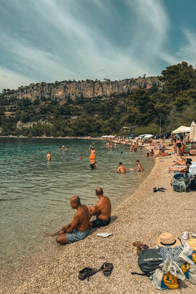 Beach goers lounging along a crescent shaped beach in Split with cliffs and trees in the background.
