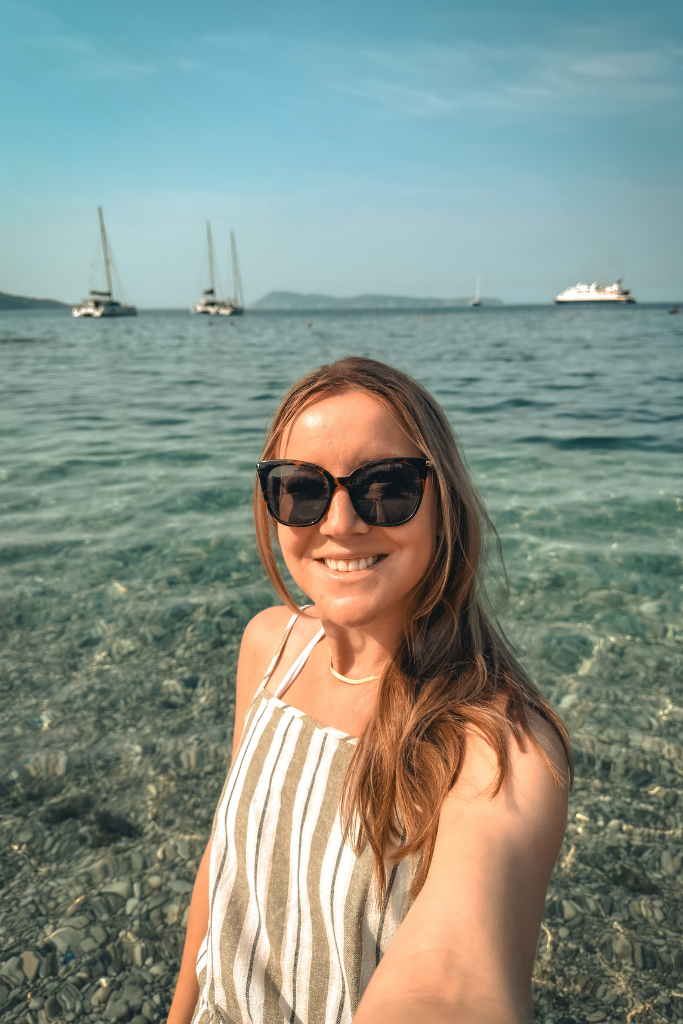 Kate taking a selfie standing on a sea shore with sailboats and turquoise water behind her.