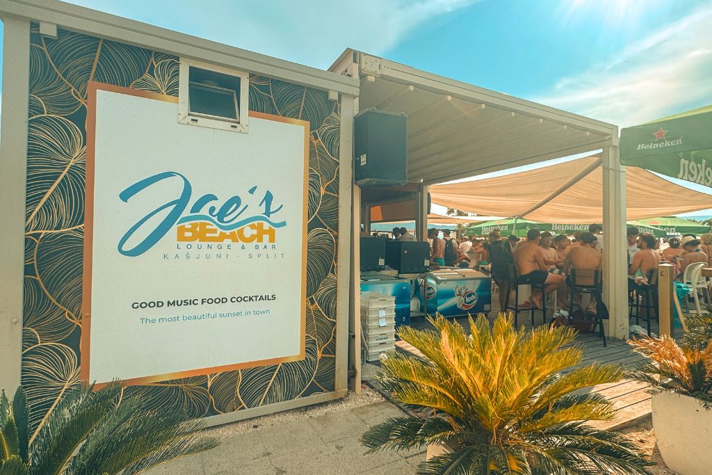 Large sign for Joes Beach Bar with a covered patio full of tables. All full of beach goers enjoying a sunny day at the beach.