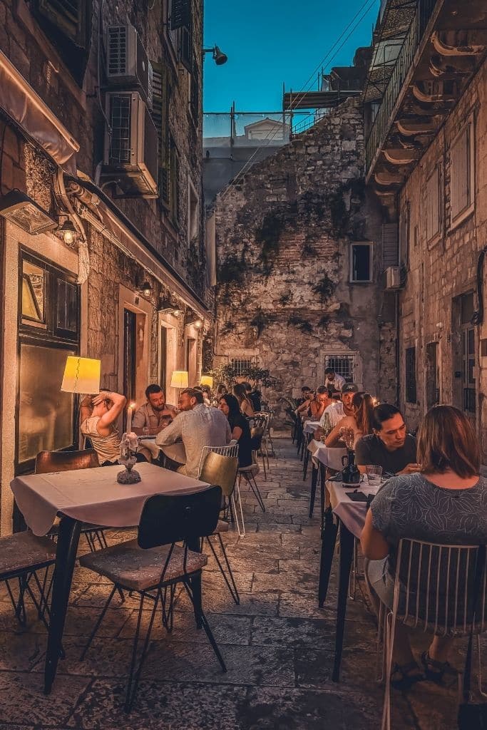 Dining tables with white table cloths line a narrow cobble stone street with people eating under candle light.