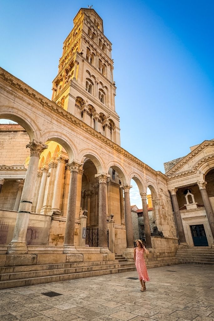 Kate stands in the courtyard of Diocletian's Palace beneath the towering Bell Tower of Saint Domnius, surrounded by grand Roman columns and historic stone architecture.