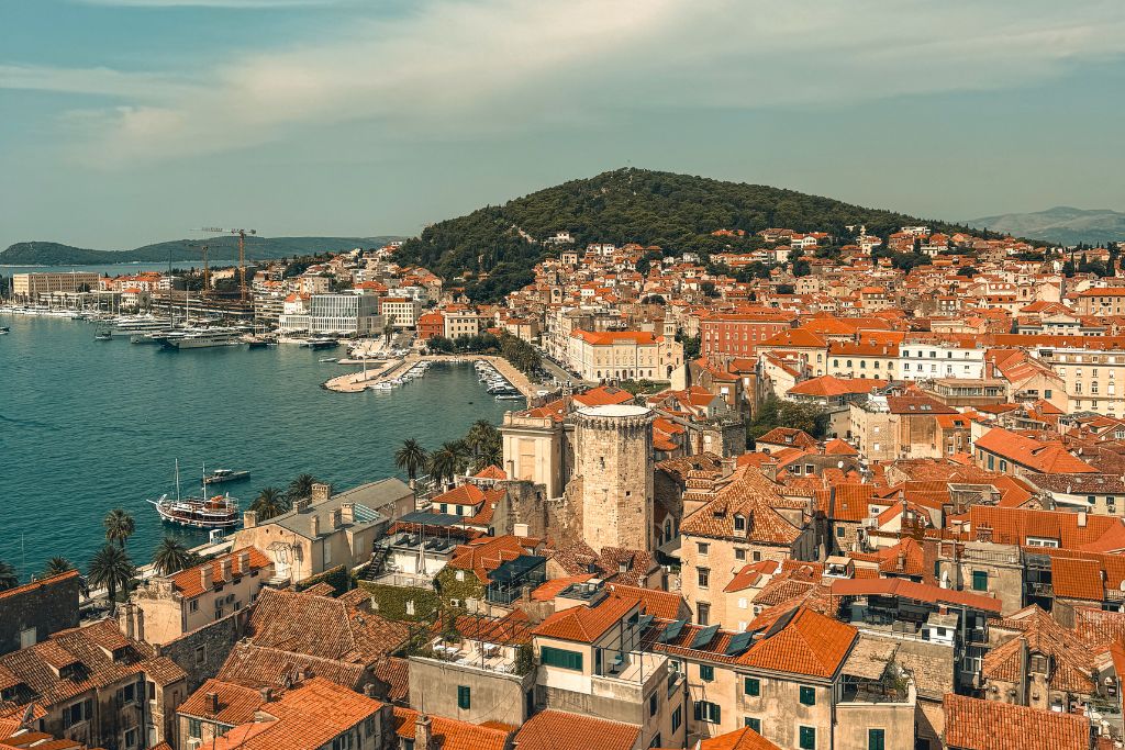 A panoramic view over Split's Old Town shows a sea of orange-tiled rooftops, the harbor filled with boats, and Marjan Hill rising in the background.