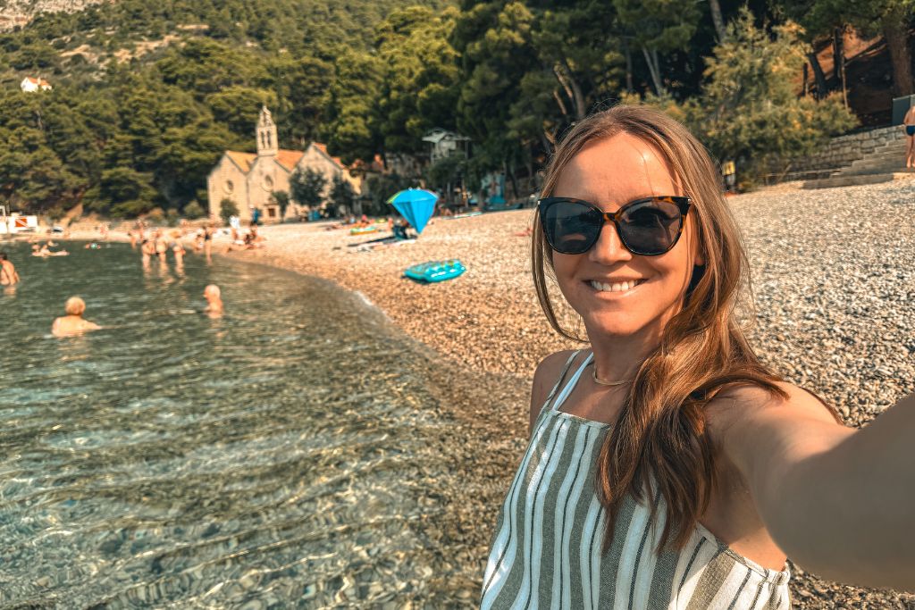 Kate takes a selfie on a beach in Vis, standing beside the crystal-clear Adriatic Sea with a pebble shoreline, swimmers, and a small stone church nestled among the trees in the background.