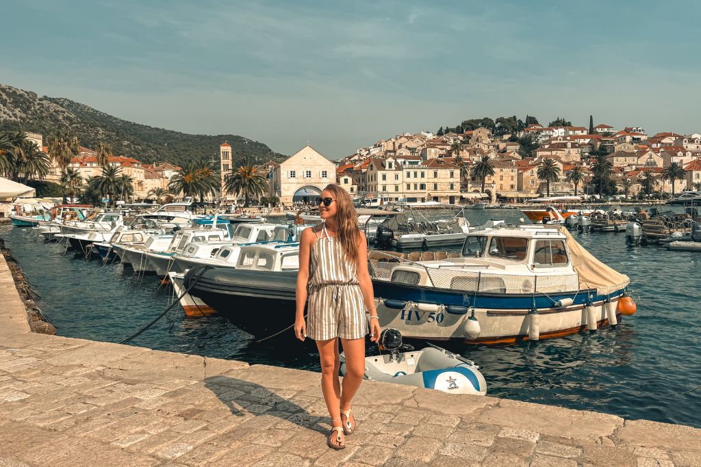 Kate stands along the waterfront in Hvar, surrounded by fishing boats and a picturesque harbor lined with stone buildings, palm trees, and red-roofed houses climbing the hillside behind her.