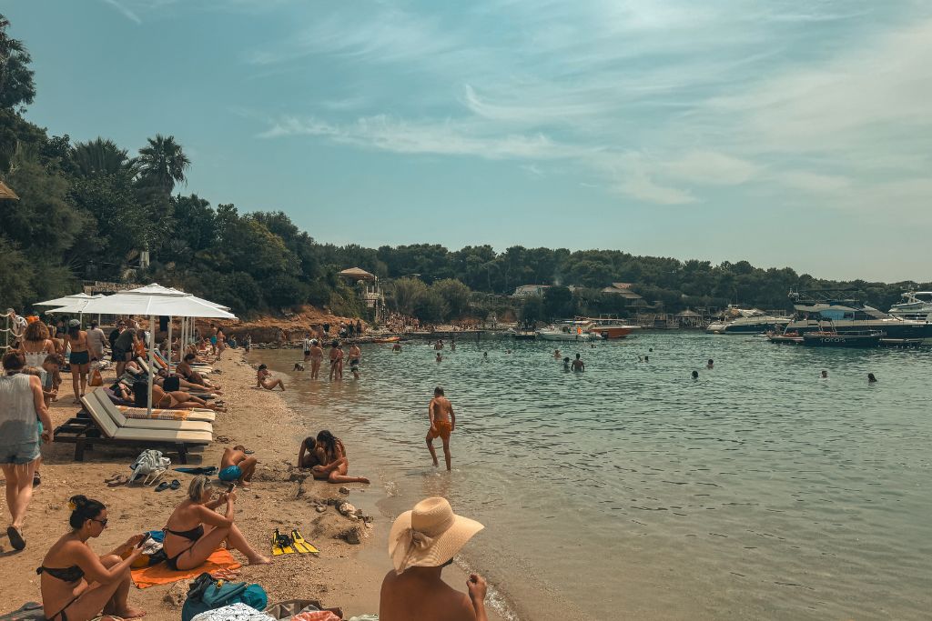 A busy beach along the Adriatic coast is filled with swimmers, sunbathers, and boats anchored in a calm bay surrounded by pine trees and waterfront cafés.