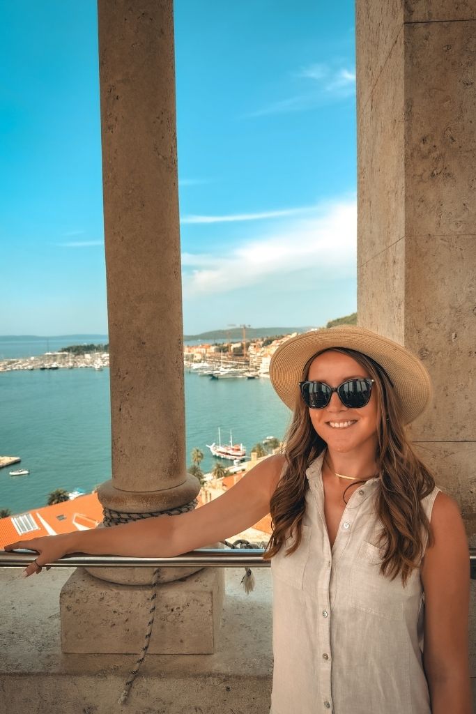 Kate stands inside the Bell Tower of Saint Domnius, smiling beside the stone columns with sweeping views of Split's harbor, red rooftops, and the Adriatic Sea visible in the background.