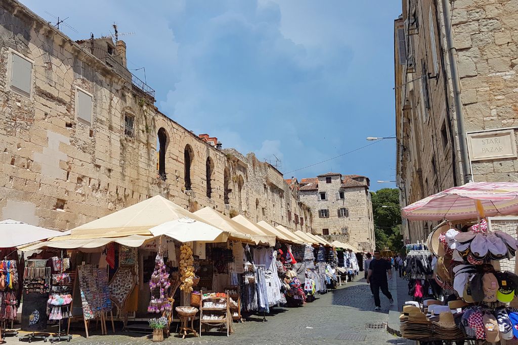 A row of market stalls lines a narrow street beside the ancient stone walls of Diocletian's Palace, with vendors selling souvenirs, clothing, and hats in Split's Old Town.