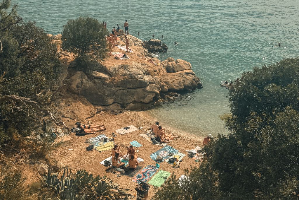 A hidden cove near Kašjuni Beach is dotted with sunbathers relaxing on towels, while swimmers gather on the rocky outcrop and enjoy the clear turquoise waters below.