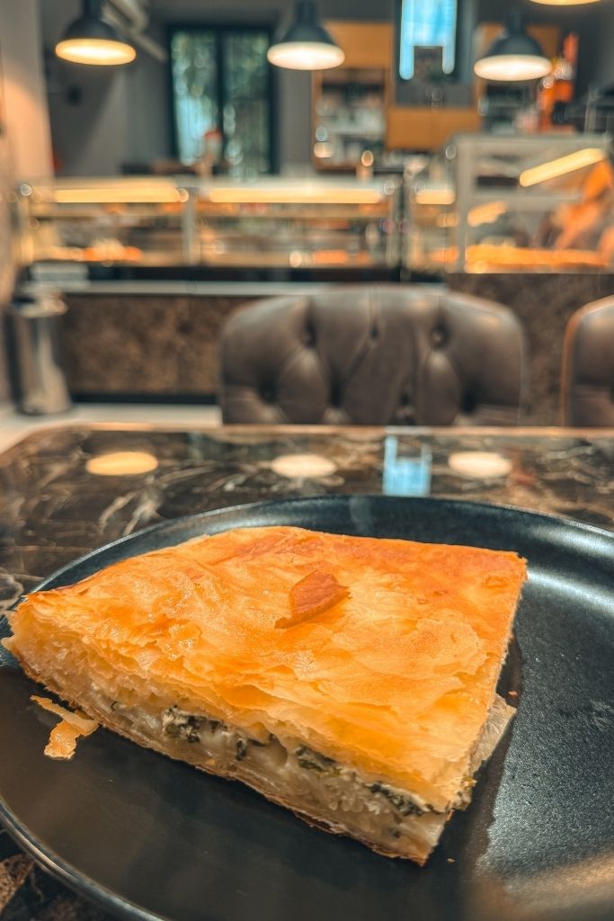 A slice of traditional Croatian burek sits on a plate inside a local bakery, showcasing its flaky golden pastry layers and savory filling.