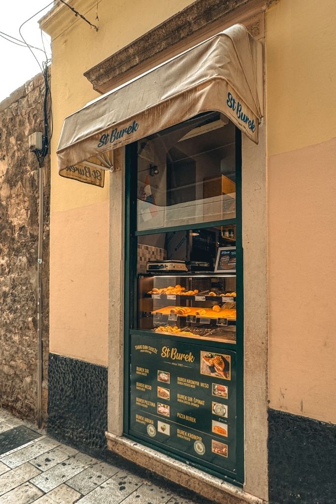 A small takeaway bakery tucked along a narrow street in Split displays freshly baked burek in its window, offering a quick and popular local snack for visitors and residents alike.