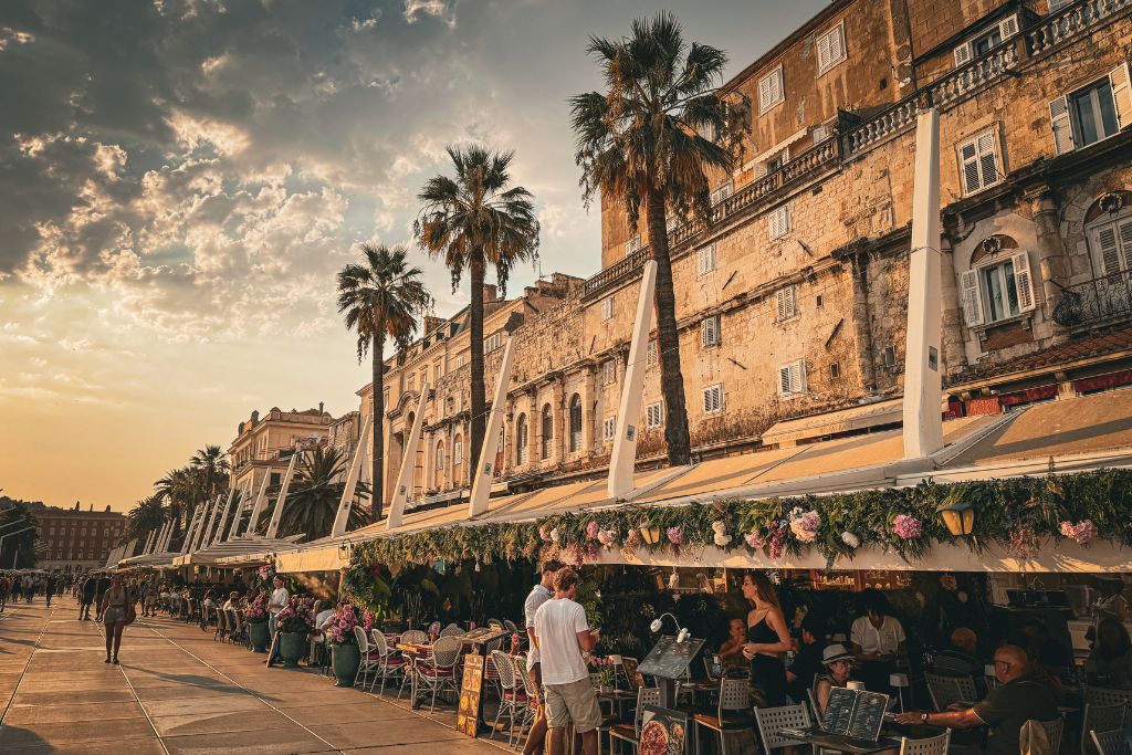 The Riva waterfront comes alive at sunset as people stroll beneath towering palm trees and dine at outdoor cafés overlooking Split's historic harbor and Adriatic coastline.