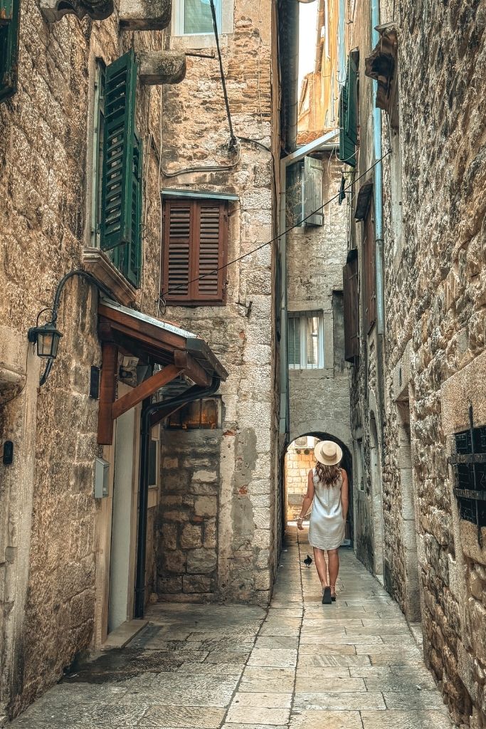 Kate strolls through one of Split's narrow stone alleyways, where weathered buildings, shuttered windows, and centuries-old architecture create the timeless atmosphere of the Old Town.