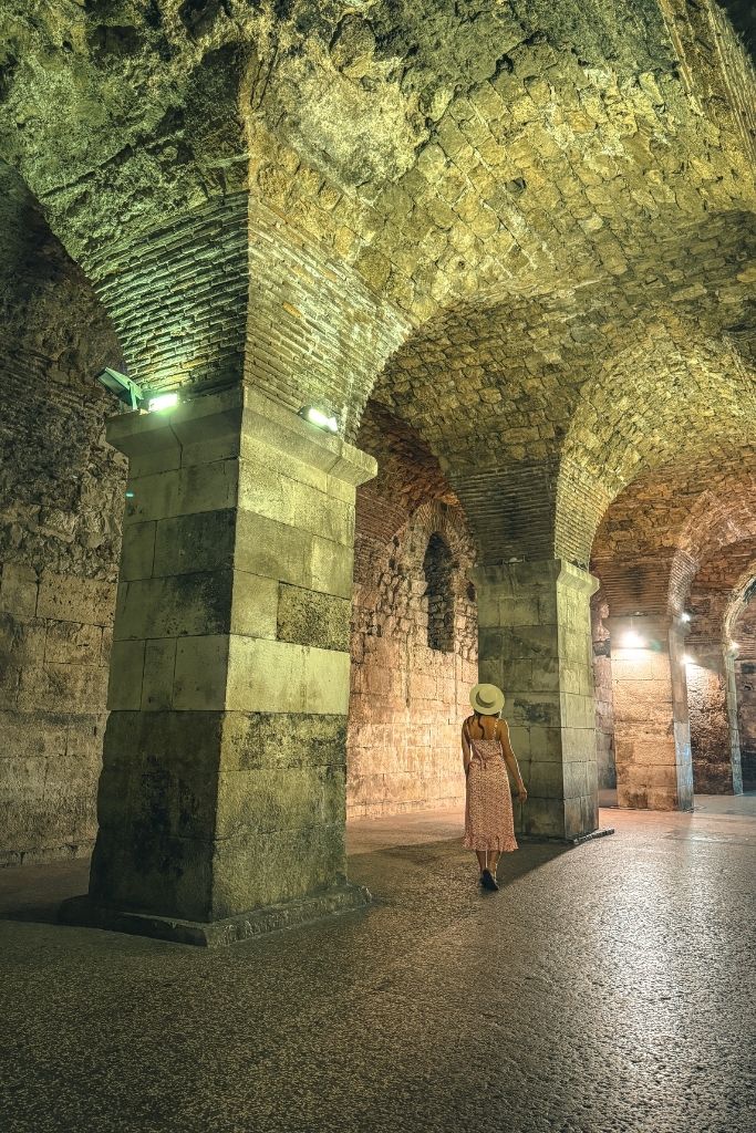 Kate walks through the underground cellars of Diocletian's Palace, surrounded by massive stone columns, vaulted brick ceilings, and ancient Roman architecture.