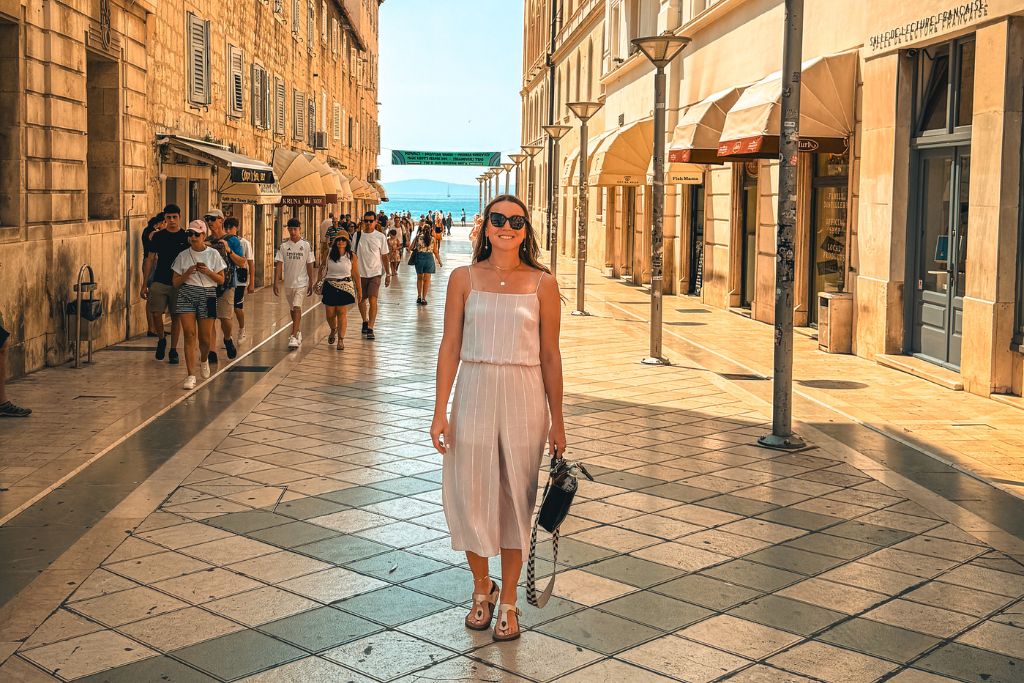 Kate stands on a bustling pedestrian street in Split's city center, surrounded by historic stone buildings, shops, and cafés, with the Adriatic Sea visible at the end of the street.