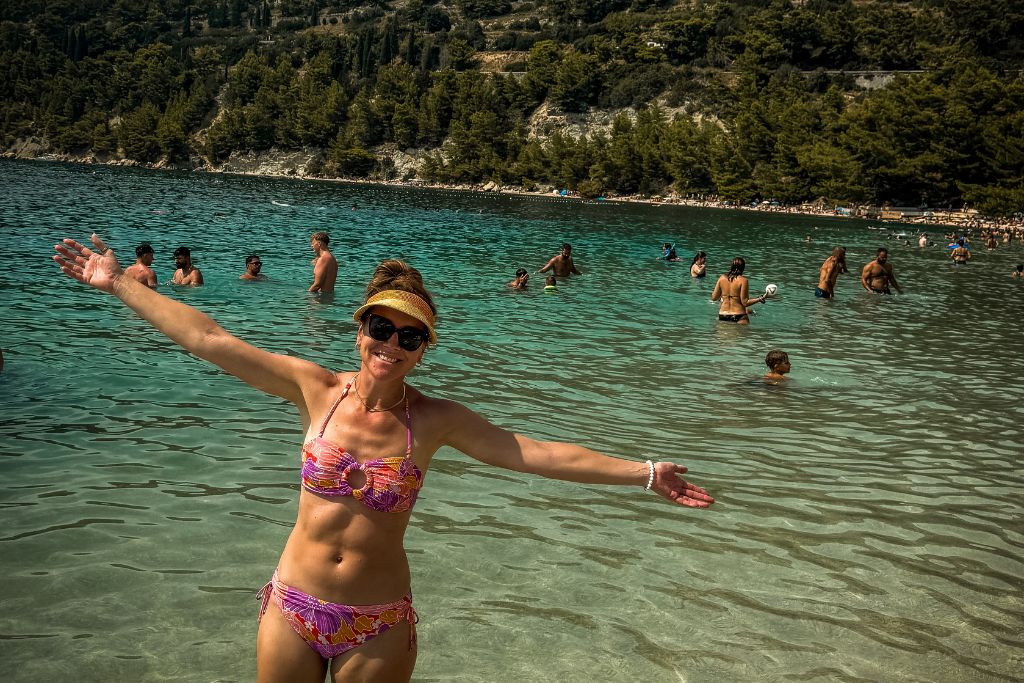 Kate stands in the crystal-clear waters of Kašjuni Beach with her arms outstretched, enjoying a sunny day on the Adriatic coast while swimmers relax in the bay behind her.