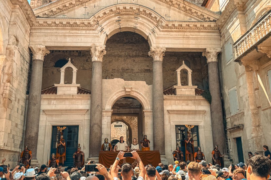 A crowd gathers in the Peristyle Square of Diocletian's Palace to watch the daily Changing of the Roman Guard ceremony, where actors portraying Emperor Diocletian, the Empress, and Roman soldiers stand beneath towering ancient columns.