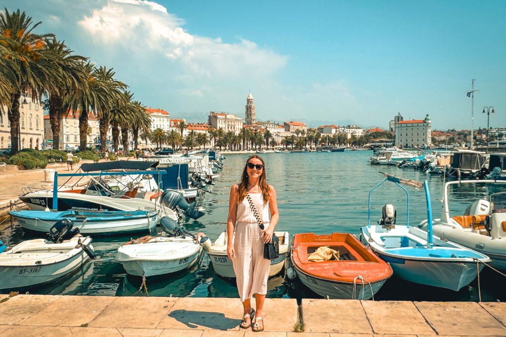 Kate stands along Split's waterfront marina with colorful boats in the foreground, palm trees lining the promenade behind her, and the historic skyline of Split stretching along the harbor in the background.