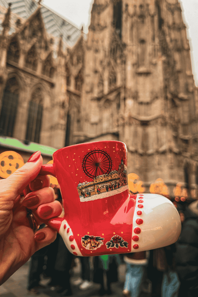 A hand holds a red boot-shaped Christmas market mug in front of St. Stephen’s Cathedral in Vienna.