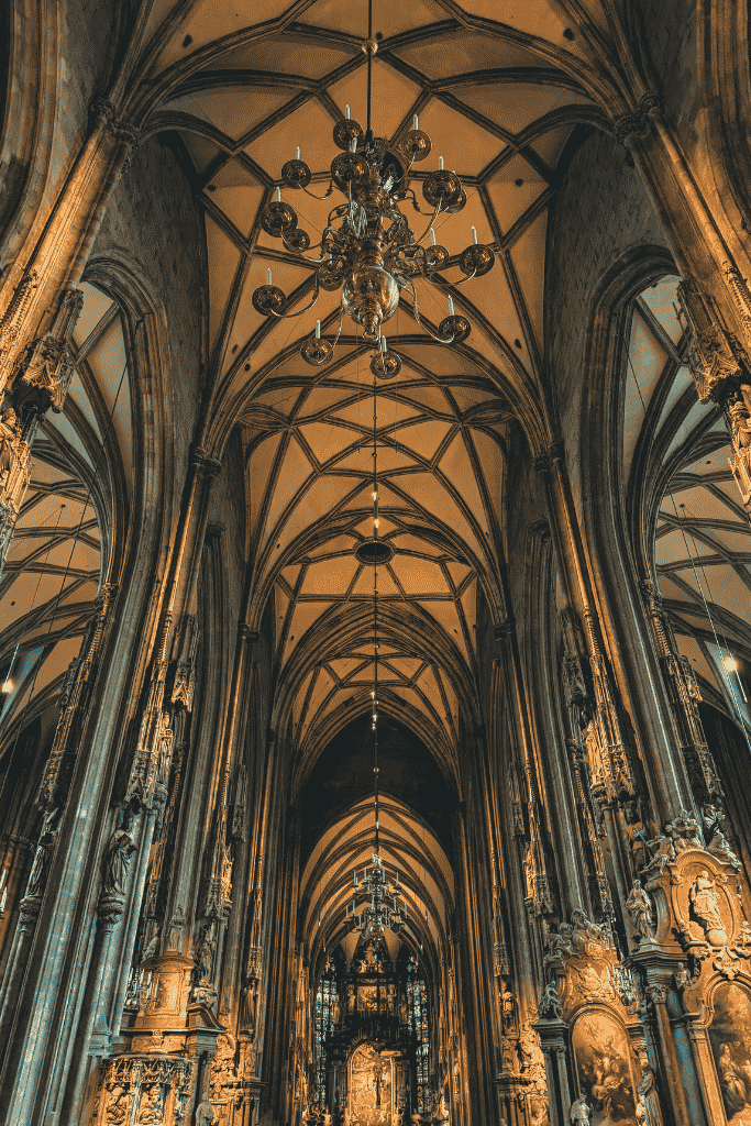 The interior of St. Stephen’s Cathedral in Vienna has tall stone columns, a high arched ceiling, and large hanging chandeliers.