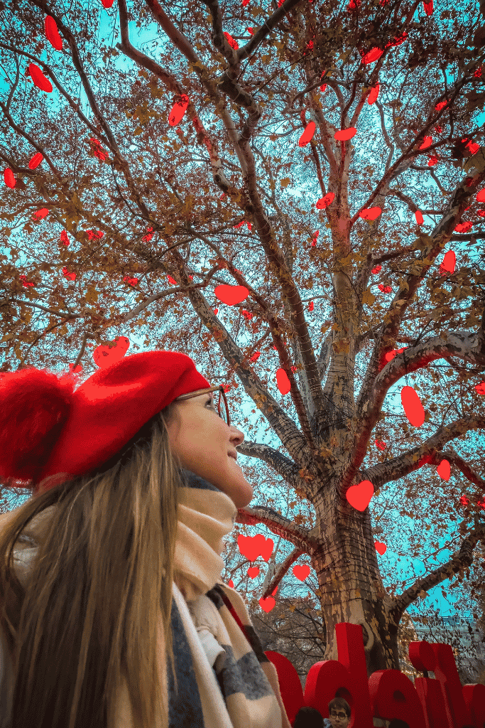 Kate in a red beret looks up at a large tree decorated with bright red heart-shaped ornaments.