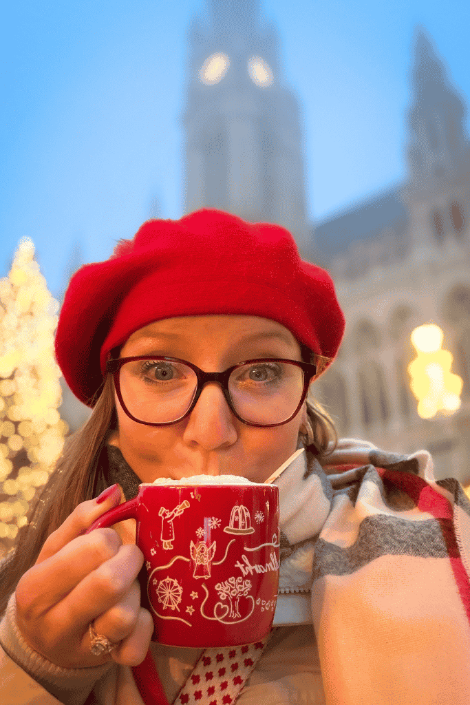 Kate in a red beret and glasses drinks from a red Christmas mug at a festive market in Vienna.
