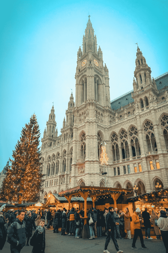 Vienna City Hall rises behind a large lit Christmas tree and busy wooden market stalls filled with visitors.