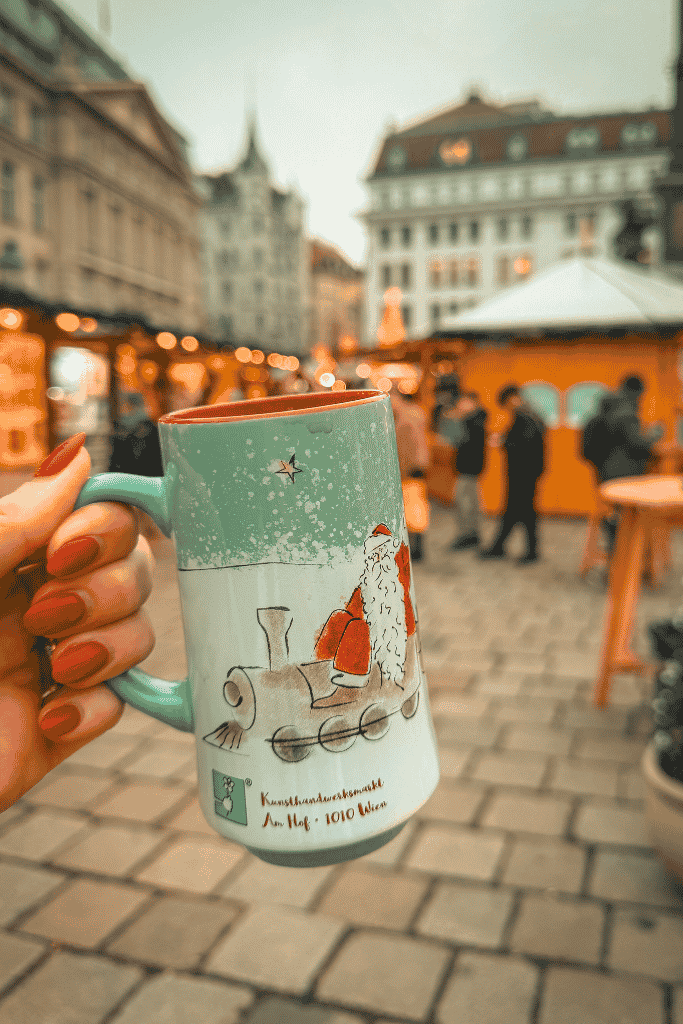 A hand holds a light green Christmas market mug decorated with Santa riding a train, with glowing market stalls in the background.
