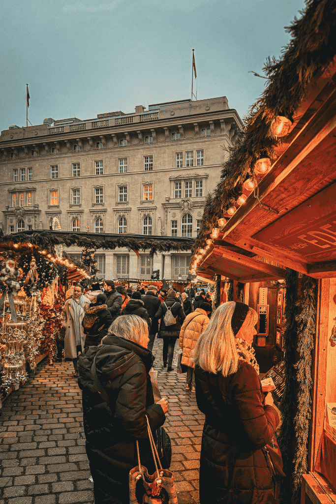 A busy Christmas market fills a cobblestone square with wooden stalls, glowing lights, and crowds of people.