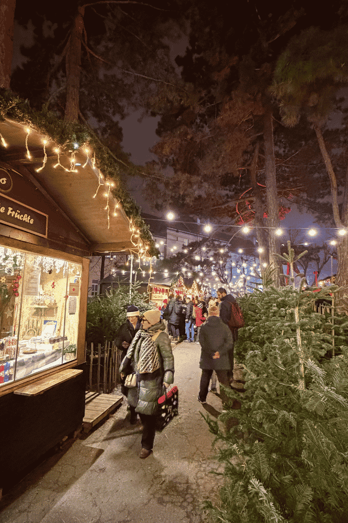 A Christmas market at night glows with string lights as people walk between wooden stalls and rows of evergreen trees.