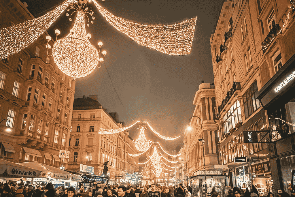 A crowded city street at night glows under large golden Christmas lights hanging between historic buildings.