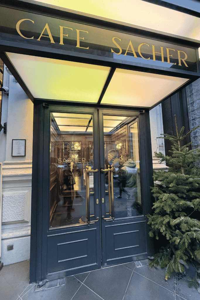 The entrance to Café Sacher has dark double doors, a glowing canopy, and a small evergreen tree beside it.