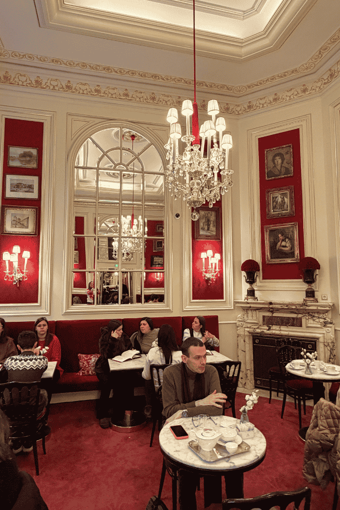 An elegant café dining room has red walls and seating, crystal chandeliers, large mirrors, framed artwork, and guests sitting at marble tables.