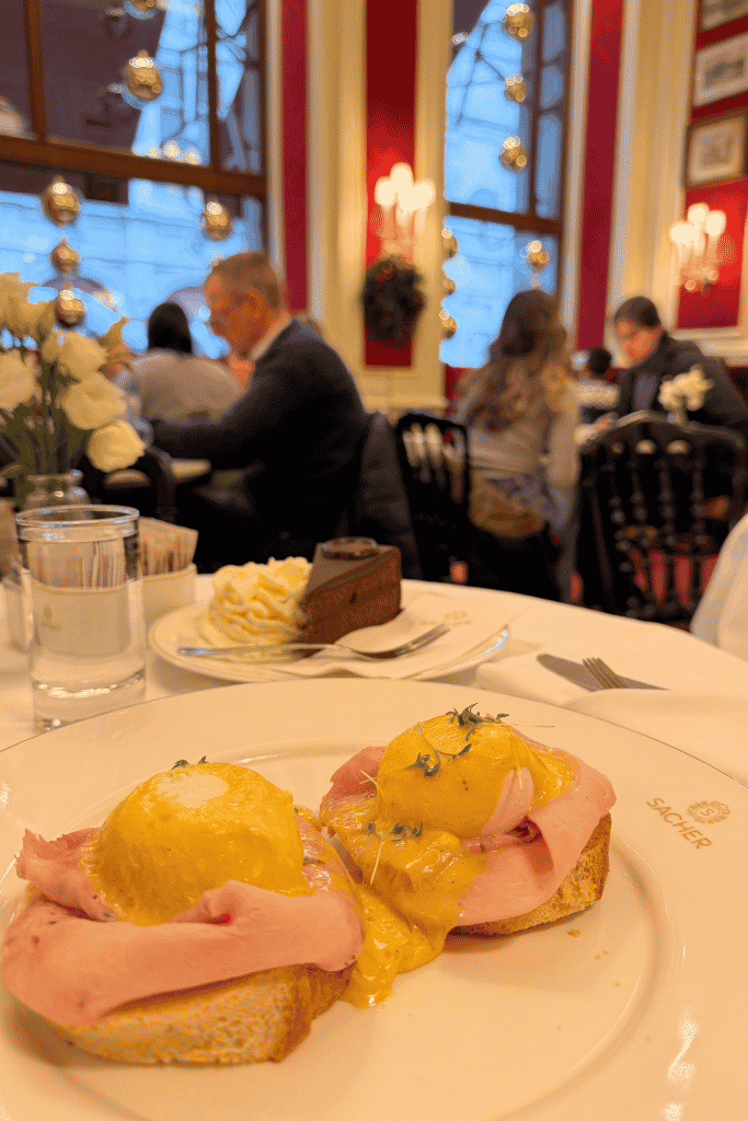 Eggs Benedict with ham and yellow sauce is served at Café Sacher, with a slice of chocolate cake and whipped cream in the background.